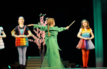 Zdjęcie z przedstawienia teatralnego, w którym grają dzieci. Na pierwszym planie dziewczynka odwrócona tyłem. Ma ubraną długą zieloną sukienkę i macha różdżką. Włosy ma upięte w warkocz. Na drugim planie rozmyte postaci innych dziewczynek, które są ubrane w kolorowe, tęczowe spódnice.