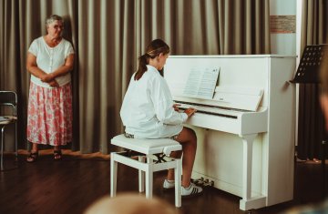 Na zdjęciu kobieta gra na pianinie. Jest odwrócona tyłem do obiektywu. Pianino ma kolor biały.  Po lewej stronie stoi nauczycielka i wpatruje się w uczennicę