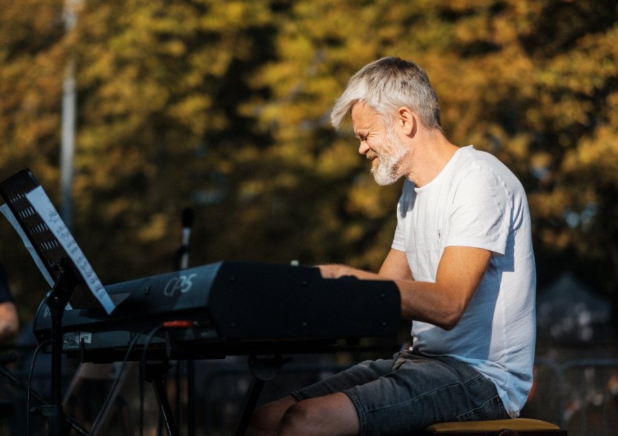 Mężczyzna grający na keyboardzie. Ma siwe włosy i siwą brodę. W tle drzewa, widać liście w kolorze żółtym. Zdjęcie wykonane podczas jesiennego pikniku w parku.
