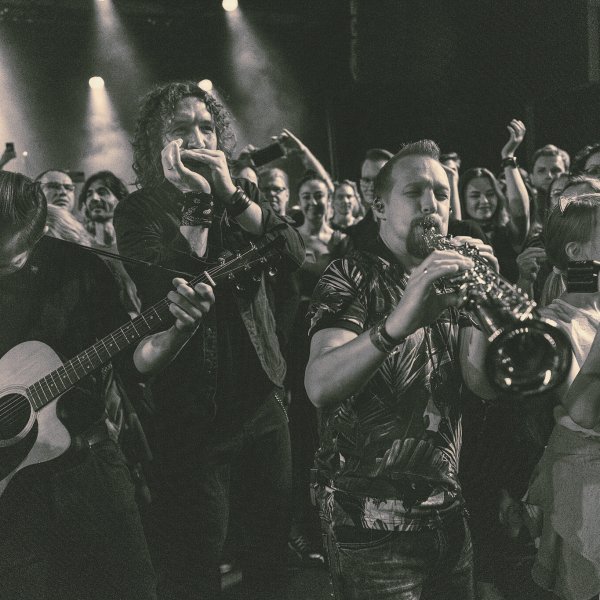 Czarno-białe zdjęcie koncertowe zespołu Kraków Street Band grającego w tłumie ludzi. Na pierwszym planie muzyk grający na gitarze akustycznej, obok niego harmonijkarz i saksofonista, a wśród publiczności znajduje się również dziewczynka trzymająca kamerę. Tłum ludzi wokół słucha, nagrywa i cieszy się muzyką na żywo.