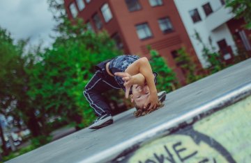 Chłopak tańczy breakdance. W tle bloki mieszkalne. Jego figura to wykonanie mostka a jedną ręką pokazuje coś do obiektywu. Ma granatowe ubranie, podłoże jest betonowe