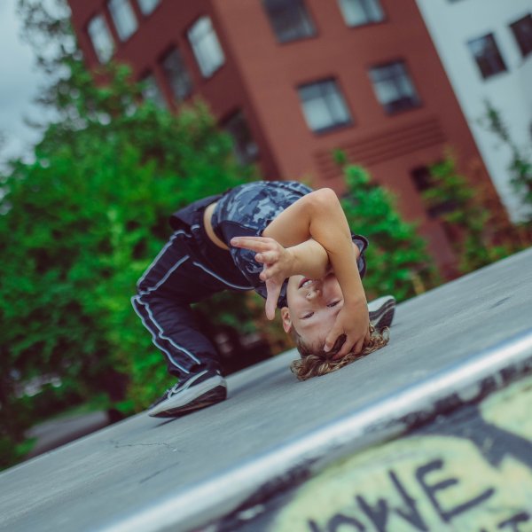 Chłopak tańczy breakdance. W tle bloki mieszkalne. Jego figura to wykonanie mostka a jedną ręką pokazuje coś do obiektywu. Ma granatowe ubranie, podłoże jest betonowe