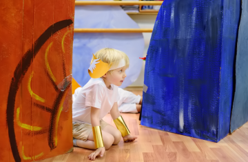 Dziewczynka w trakcie przedstawienia teatralnego. Dziewczynka to dziecko, które ma około 3 lata. Klęczy z rękami opartymi na podłodze między dwoma blokami scenografii. Po prawej stronie blok ma kolor niebieski po lewej pomarańczowy. Na głowie dziewczynka ma opaskę z żółtym daszkiem