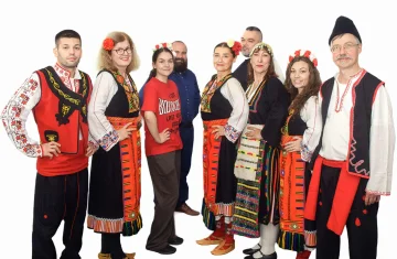 Na zdjęciu grupa folklorystyczna Bulgarica podczas występu. 5 osób ubranych w ludowe bułgarskie stroje. Po prawej i po lewej mężczyzna, a w środku trzy kobiety. Grupa trzyma się za ręce i tańczy podnosząc lewą nogę, .