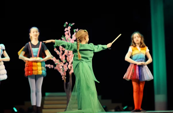 Zdjęcie z przedstawienia teatralnego, w którym grają dzieci. Na pierwszym planie dziewczynka odwrócona tyłem. Ma ubraną długą zieloną sukienkę i macha różdżką. Włosy ma upięte w warkocz. Na drugim planie rozmyte postaci innych dziewczynek, które są ubrane w kolorowe, tęczowe spódnice.