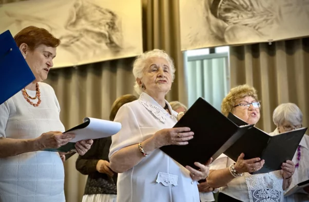 grupa seniorek śpiewa, kobiety ubrane są w białe koszule, w ręku trzymają tekst z utworami