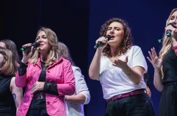 Grupa kobiet śpiewa do mikrofonu. Grupa ubrana jest w różowo białe stroje