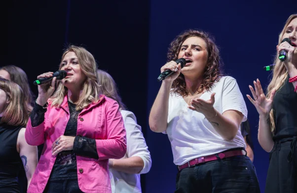 Grupa kobiet śpiewa do mikrofonu. Grupa ubrana jest w różowo białe stroje
