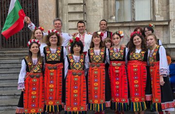 Na zdjęciu grupa folklorystyczna Bulgarica podczas występu. 5 osób ubranych w ludowe bułgarskie stroje. Po prawej i po lewej mężczyzna, a w środku trzy kobiety. Grupa trzyma się za ręce i tańczy podnosząc lewą nogę, .