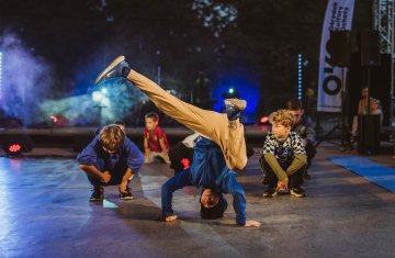 Grupa dzieci trenuje breakdance na scenie – na pierwszym planie chłopiec w niebieskiej bluzie wykonuje dynamiczną figurę na głowie, a wokół niego pozostali uczestnicy obserwują lub przygotowują się do ruchu. W tle kolorowe światła sceniczne i widownia.