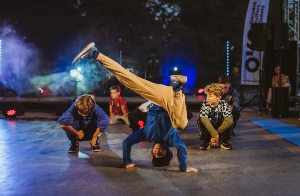 Grupa dzieci trenuje breakdance na scenie – na pierwszym planie chłopiec w niebieskiej bluzie wykonuje dynamiczną figurę na głowie, a wokół niego pozostali uczestnicy obserwują lub przygotowują się do ruchu. W tle kolorowe światła sceniczne i widownia.