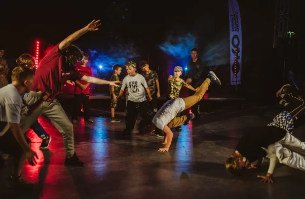 grupa dzieci tańczy na boisku taniec breakdance. Jest ciemno i oświetlają ich czerwono niebieskie światła
