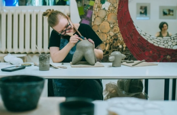 Kobieta siedzi przy stole i pracuje nad glinianą figurą.