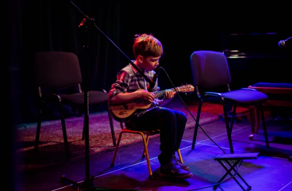 Na scenie siedzi chłopiec i gra na ukulele