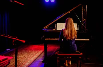 Na scenie przy fortepianie tyłem siedzi kobieta.