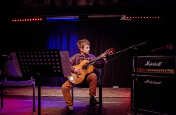 Na scenie, na krześle siedzi chłopiec i gra na gitarze