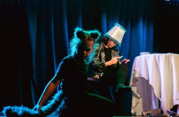Grupa pięciu dzieci stoi na scenie teatralnej w szeregu, trzymając się za ręce i uśmiechając po zakończonym występie. Dzieci mają na sobie kolorowe, teatralne kostiumy: jedno z nich nosi srebrną pelerynę i dużą, puszystą perukę, inne mają tiulowe spódnice, korony, fantazyjne nakrycia głowy i ciemne ubrania sceniczne. Tło jest ciemne, oświetlone reflektorami w odcieniach fioletu i niebieskiego.