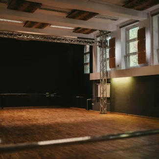 Zdjęcie sali widowiskowej Ośrodka Kultury Ochoty. Na pierwszym planie parkiet drewniany a na drugim scena koloru czarnego. Jest również czarne okotarowanie.