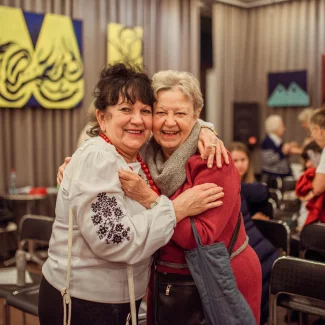 Dwie uśmiechnięte seniorki obejmują się serdecznie podczas spotkania w Klubie Kultury Seniora. W tle widać inne osoby rozmawiające i kolorowe obrazy na ścianach sali. Atmosfera ciepła, przyjaźni i wspólnoty.