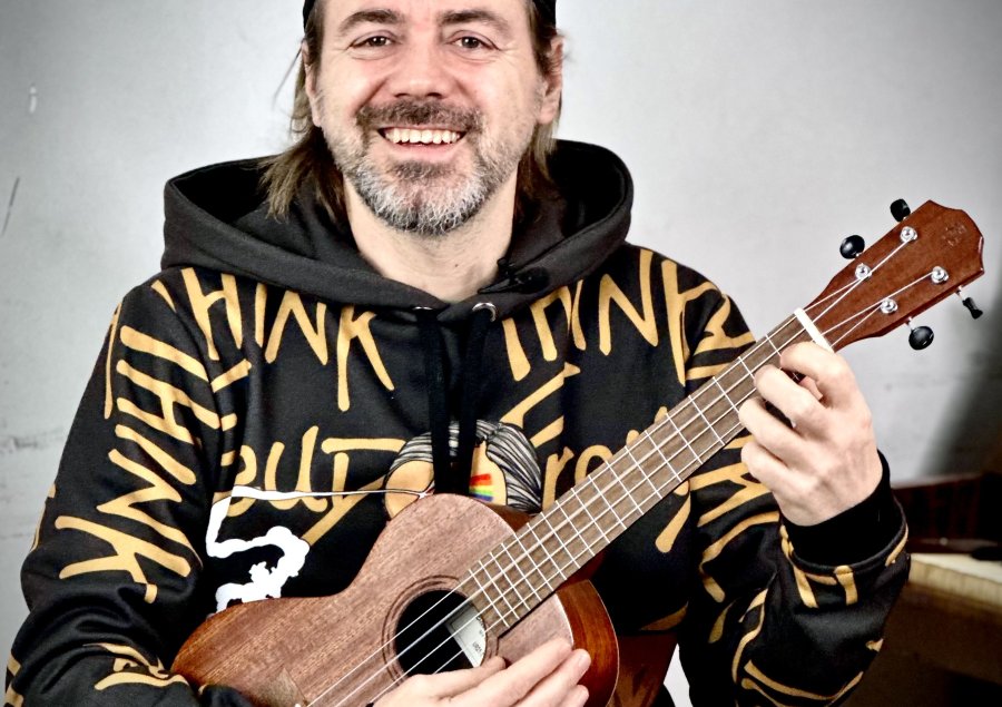 Uśmiechnięty mężczyzna w kolorowej bluzie i z czapką z daszkiem na głowie. Czapka odwrócona jest daszkiem do tyłu. Gra na ukulele. Ma siwiejącą brodę