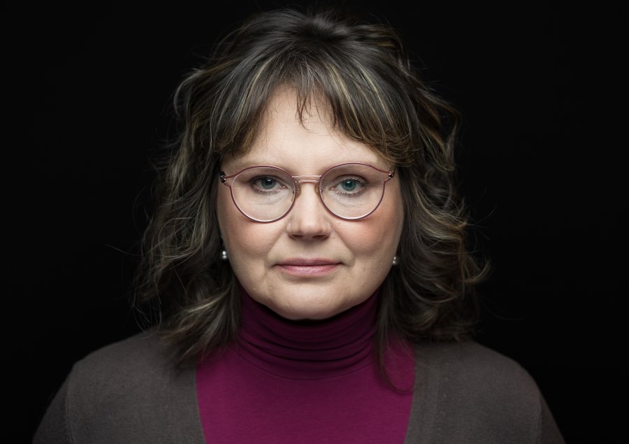 Zdjęcie portretowe kobiety w okularach na czarnym tle. Ma ciemne włosy i grzywkę, lekko się uśmiecha.