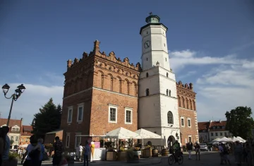 Ratusz w Sandomierzu – zabytkowy, ceglasty budynek z jasną wieżą zegarową na rynku, widoczny w słoneczny dzień, z ludźmi spacerującymi wokół.