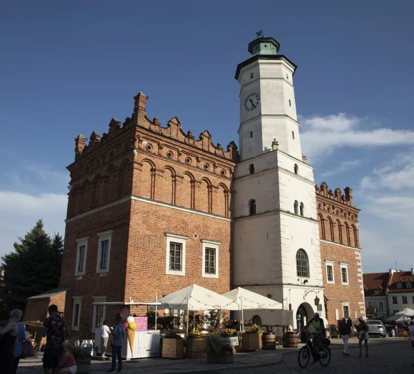 Ratusz w Sandomierzu – zabytkowy, ceglasty budynek z jasną wieżą zegarową na rynku, widoczny w słoneczny dzień, z ludźmi spacerującymi wokół.