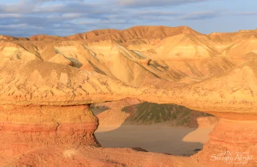 In the photo, there is a desert. In the center of the image, there is something, but we cannot see what it is; it casts a large shadow on the sand.