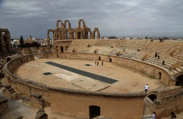 Rzymski amfiteatr z kamiennymi trybunami i ruinami łuków, widok z góry na arenę pod pochmurnym niebem.