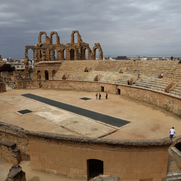 Rzymski amfiteatr z kamiennymi trybunami i ruinami łuków, widok z góry na arenę pod pochmurnym niebem.
