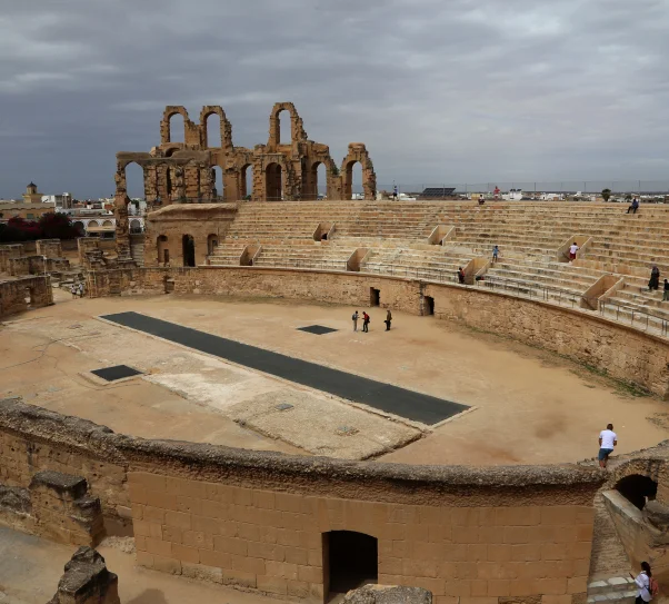 Rzymski amfiteatr z kamiennymi trybunami i ruinami łuków, widok z góry na arenę pod pochmurnym niebem.