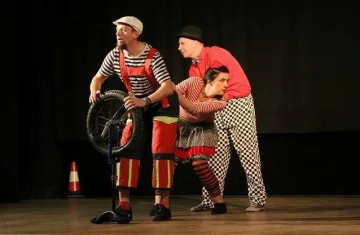 Trójka artystów cyrkowych w kolorowych, klaunowskich kostiumach występuje na scenie. Jeden z nich trzyma koło od monocykla, drugi stoi za nim w czerwonej koszuli i czarno-białych spodniach w kratę, a trzeci przytula się do niego z teatralną miną. W tle ciemna kurtyna i pachołek ostrzegawczy. Scena oświetlona światłem scenicznym.