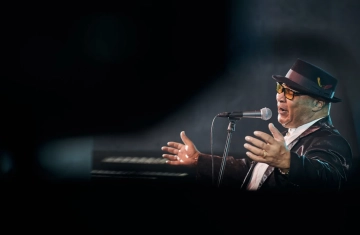 Czarny mężczyzna w kapeluszu śpiewa do mikrofonu. Jest ustawiony bokiem do obiektywu. Ma czarby kapelusz i czarny garnitur. Tło ciemne, zadymione. Zdjęcie wykonane podczas koncertu.