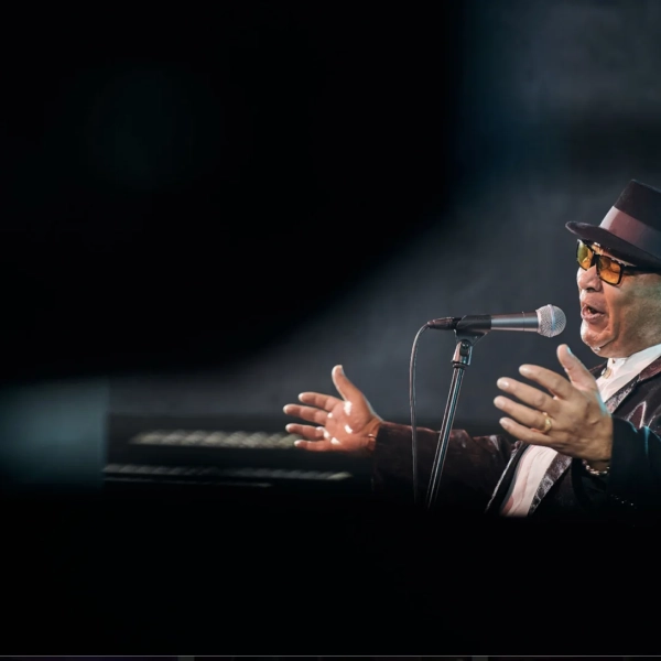 Czarny mężczyzna w kapeluszu śpiewa do mikrofonu. Jest ustawiony bokiem do obiektywu. Ma czarby kapelusz i czarny garnitur. Tło ciemne, zadymione. Zdjęcie wykonane podczas koncertu.