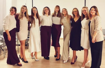 Grupa dziewczyn w stronach w odcieniach beżu i brązu. Stoją przytulając się od tyłu w półkolu.