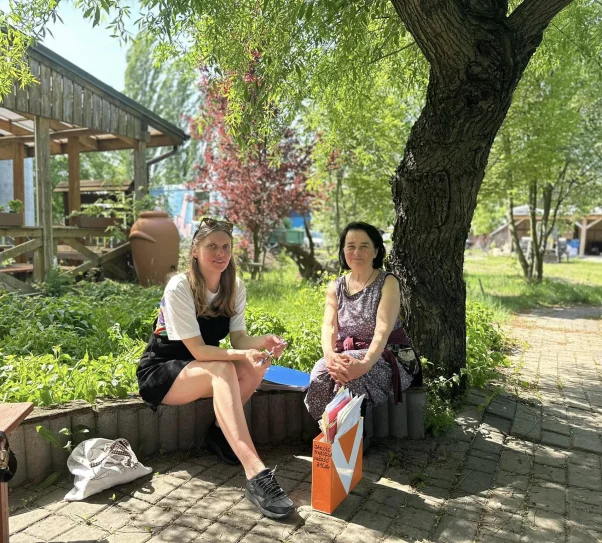 Dwie kobiety siedzą w cieniu drzewa w zielonym ogrodzie. Rozmawiają, otoczone roślinnością i drewnianymi zabudowaniami. Na pierwszym planie widać teczkę z dokumentami i letnie światło przenikające przez liście.