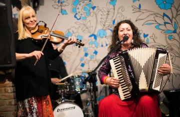 zdjęcie przedstawia dwie kobiety. Jedna stoi i gra na skrzypcach, druga siedzi i gra na akordeonie