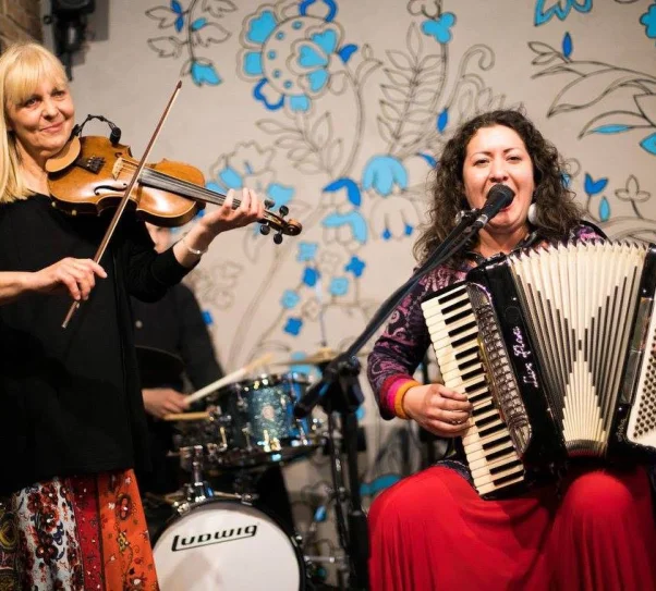 zdjęcie przedstawia dwie kobiety. Jedna stoi i gra na skrzypcach, druga siedzi i gra na akordeonie