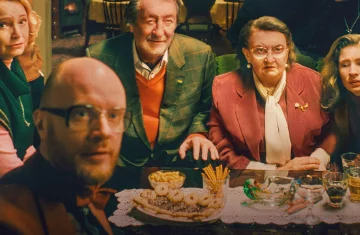 Na zdjęciu widzimy grupę sześciu osób siedzących przy stole, wyglądających prosto w obiektyw, jakby właśnie przerwano im rozmowę lub ktoś niespodziewanie zrobił im zdjęcie. Atmosfera przypomina rodzinne spotkanie — być może święta lub uroczysty obiad. Stół przykryty jest koronkowym bieżnikiem i zastawiony talerzem z ciasteczkami, paluszkami, orzeszkami oraz kilkoma szklankami z napojami.  Postacie są różnorodne wiekowo — starszy mężczyzna w marynarce i swetrze w centrum, obok niego starsza kobieta w bordowym żakiecie z wyrazem powagi na twarzy, młodsza kobieta z długimi blond włosami oraz inni uczestnicy o zróżnicowanych wyrazach twarzy: zdziwienie, zaintrygowanie, dezorientacja. Ujęcie ma ciepłą kolorystykę i przytulny, nostalgiczny klimat, przypominający sceny z europejskich filmów obyczajowych.