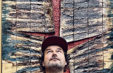 A bearded man wearing a black T-shirt with colorful illustrations and a red cap stands in front of a large textured art installation in shades of brown, red, and green. He is looking upward, appearing thoughtful or intrigued.