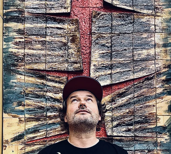 A bearded man wearing a black T-shirt with colorful illustrations and a red cap stands in front of a large textured art installation in shades of brown, red, and green. He is looking upward, appearing thoughtful or intrigued.