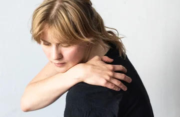 Zdjęcie przedstawia kobietę, którą widać do połowy, stoi bokiem z głową pochyloną z zamkniętymi oczami, prawą reką trzyma się lewego barku