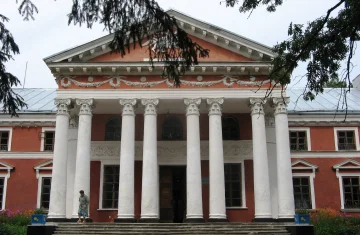 Zdjęcie przedstawia okazały, historyczny budynek w stylu klasycystycznym z czerwonymi ścianami i dużym portykiem wspartym na ośmiu białych kolumnach. Na schodach przed wejściem stoi jedna osoba, a wokół budowli rosną kwiaty i drzewa, których gałęzie częściowo wchodzą w kadr.