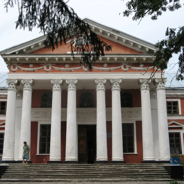 Zdjęcie przedstawia okazały, historyczny budynek w stylu klasycystycznym z czerwonymi ścianami i dużym portykiem wspartym na ośmiu białych kolumnach. Na schodach przed wejściem stoi jedna osoba, a wokół budowli rosną kwiaty i drzewa, których gałęzie częściowo wchodzą w kadr.