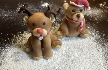 Two small Christmas-themed sugar paste figurines sit on a surface dusted with powdered sugar. On the left is a reindeer with dark antlers, a red nose, and a scarf. On the right is a bear wearing a Santa hat and a red-and-white striped scarf.