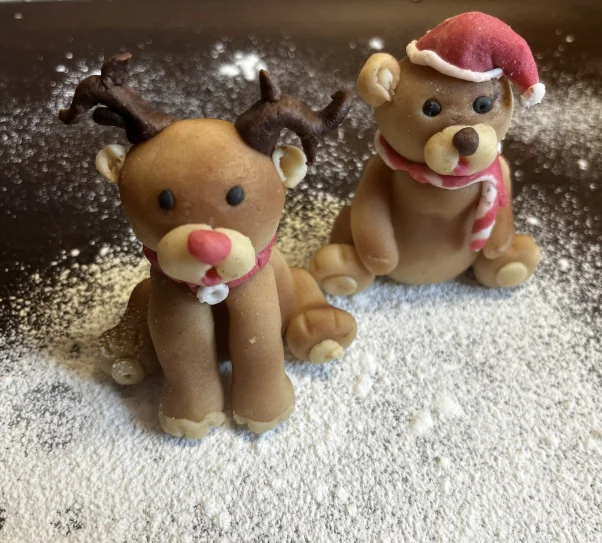 Two small Christmas-themed sugar paste figurines sit on a surface dusted with powdered sugar. On the left is a reindeer with dark antlers, a red nose, and a scarf. On the right is a bear wearing a Santa hat and a red-and-white striped scarf.