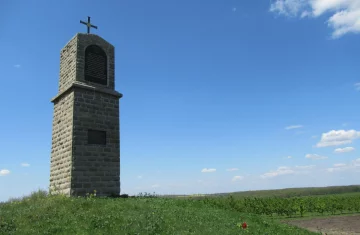 Zdjęcie przedstawia kapliczkę, która stoi na lekkim wznienieniu na trawie,  tło stanowi błękitne niebo
