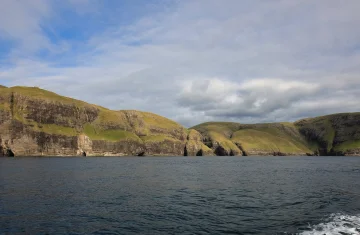 Skaliste klify Wysp Owczych porośnięte zieloną trawą, widziane od strony morza; spokojna, ciemnoniebieska woda na pierwszym planie i pochmurne, zmienne niebo nad horyzontem.