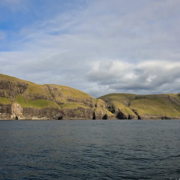 Skaliste klify Wysp Owczych porośnięte zieloną trawą, widziane od strony morza; spokojna, ciemnoniebieska woda na pierwszym planie i pochmurne, zmienne niebo nad horyzontem.