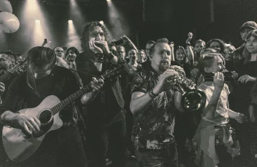 Czarno-białe zdjęcie koncertowe zespołu Kraków Street Band grającego w tłumie ludzi. Na pierwszym planie muzyk grający na gitarze akustycznej, obok niego harmonijkarz i saksofonista, a wśród publiczności znajduje się również dziewczynka trzymająca kamerę. Tłum ludzi wokół słucha, nagrywa i cieszy się muzyką na żywo.